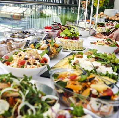 Neck down view of side of a chef in uniform and waist apron as they lay a bowl of salad on a buffet table full of bowls of cold food