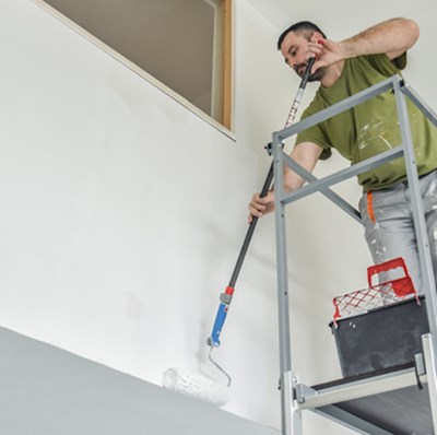 Man on a decorators scaffolding tower with a roller on an extension pole painting the wall of an intermediate floor at the top of the room