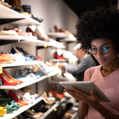 Woman looking at a digital tablet in her hands with shelves of shoes along side and behind her. 