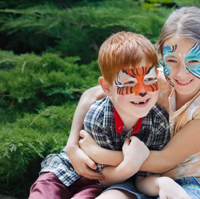 Girl with a butterfly painted on her face, her arms around a boy sitting beside her who has tiger markings painted on his, with a greenery background
