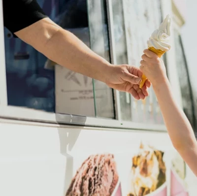 Girl reaching up to take a whipped ice cream cone from an arm coming out of the side window of an ice cream van with pictures of ice cream on the van