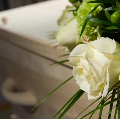 A blurred backdrop of a wooden coffin and curtains with with roses, green carnations, green berries and fern greenery in focus on top 