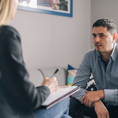 Man on a sitting on a couch talking to a women writing on paper attached to a clip board sitting on a chair opposite him Man on a sitting on a couch talking to a women writing on paper attached to a clip board sitting on a chair opposite him