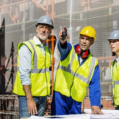 Two men and a women in hard hats and high vis, looking in the direction that one is pointing, with a backdrop of scaffolding and netting