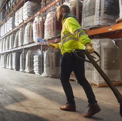 Man in a warehouse pulling a hand truck with large shelving holding large sacks, and a forklift truck in the far end of the aisle