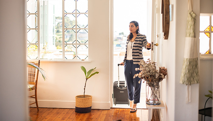 Women With Cabin Bag On Wheels Opening Door Into White Bright Room With Wooden Flooring