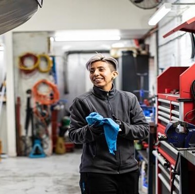 A women in a garage by a work bench with a car lifted above her to her right  A women in a garage by a work bench with a car lifted above her to her right