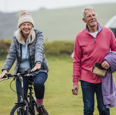 Women on a bike with a man beside her, in a field with blurred caravans, grass wall and greenery in the background