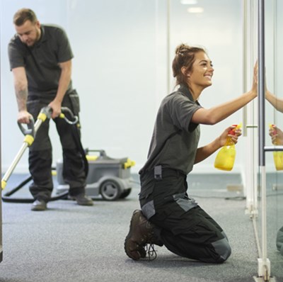 A man vacuuming an office hall by some stairs while a women is kneeling cleaning a glass walled room  A man vacuuming an office hall by some stairs while a women is kneeling cleaning a glass walled room