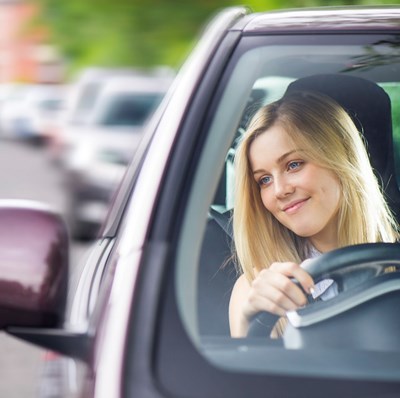 Windscreen view of a young women in the driving seat, smiling looking into her wing mirror, with a blurred queue of cars behind her
