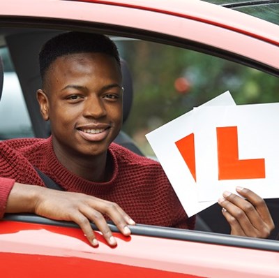 A smiling young man in the driving seat with his right arm on the open window of a car holding up two L plates 