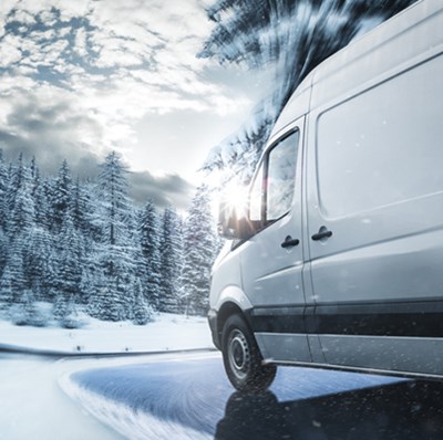 Side view of a white van taking a bend on a snowy, fir tree lined road
