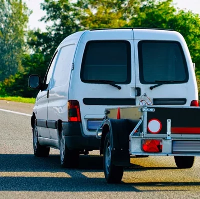 A rear view of a small van pulling a small trailer on a road with trees and hedgerows along the right hand view A rear view of a small van pulling a small trailer on a road with trees and hedgerows along the right hand view