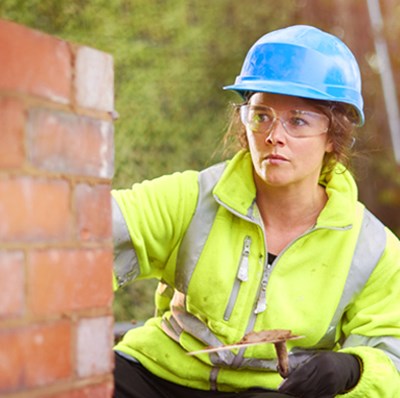 A women wearing a hard hat and high viz jacket building a wall with a spirit level in the fore ground and another builder in the blurred background