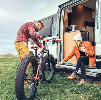 Man holding his mountain bike beside a campervan with a woman sat in the open door on the side in a field Man holding his mountain bike beside a campervan with a woman sat in the open door on the side in a field