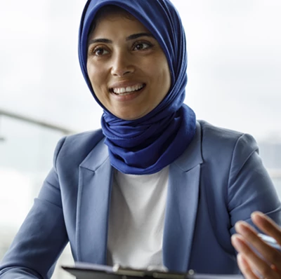 A smiling women in a head scarf and suit jacket sitting at a table with a pen and clip board on a glass balcony with a blurred back ground of tower blocks  A smiling women in a head scarf and suit jacket sitting at a table with a pen and clip board on a glass balcony with a blurred back ground of tower blocks