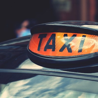 Lit taxi sign light on top of a modern London black cab, with a blurred background of some people