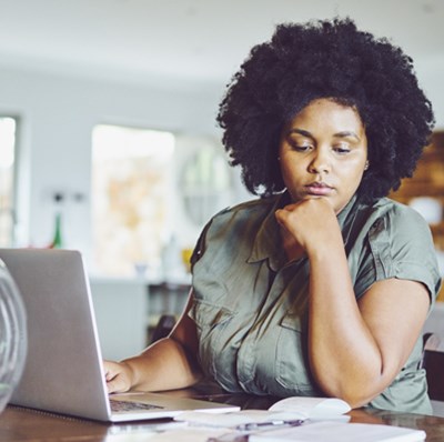 Head and shoulder view of a woman at a dining room table working on a laptop looking pensively at paperwork beside her with a blurred background  Head and shoulder view of a woman at a dining room table working on a laptop looking pensively at paperwork beside her with a blurred background