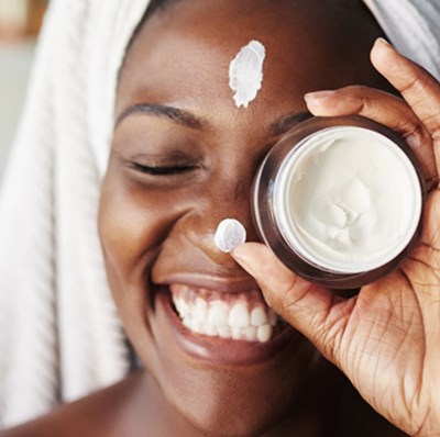 Head shot of a smiling woman with her hair in a towel holding an open jar of cream with the bottom against her eye