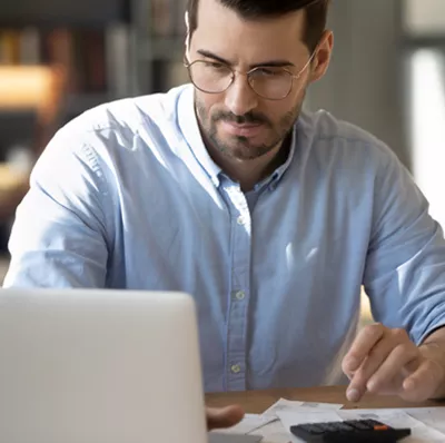 A man working on a laptop and a calculator on a desk with paper, a pot with pencils and a plant with a blurred background of a bookshelf and chair A man working on a laptop and a calculator on a desk with paper, a pot with pencils and a plant with a blurred background of a bookshelf and chair