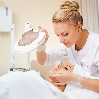 Beautician Angling Lamp At Women Lying On A Treatment Table Beautician Angling Lamp At Women Lying On A Treatment Table
