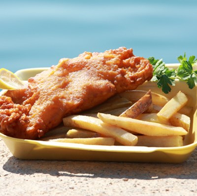A cardboard bowl of fish and chips with a slice of lemon and a side of mushy peas and lime on exposed aggregate concrete with a water background A cardboard bowl of fish and chips with a slice of lemon and a side of mushy peas and lime on exposed aggregate concrete with a water background