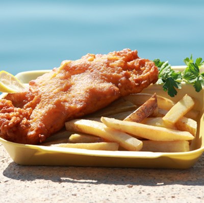 A cardboard bowl of fish and chips with a slice of lemon and a side of mushy peas and lime on exposed aggregate concrete with a water background