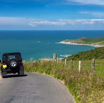 A rear view of a jeep driving down hill on a country lane towards the sea with views of grassy hills ending in bays to the water