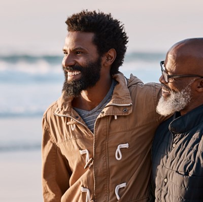 Younger man with arm around the shoulder of an older man with his arm across the youngers back, walking along the beach Younger man with arm around the shoulder of an older man with his arm across the youngers back, walking along the beach