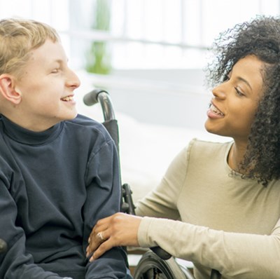 Woman crouched down to level of boy in a wheel chair, they are smiling at each other with a blurred hospital bed in a ward backdrop