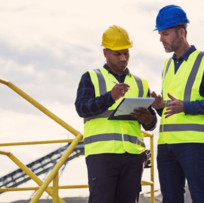 Two men in hard hats and high viz jackets, writing on paper on a clip board while the other is gesticulates, standing on top of a building by railings