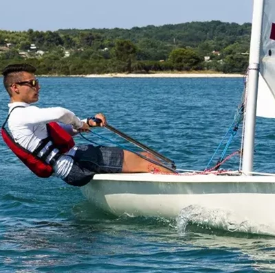 Man leaning out over the edge of the side of a small boat holding on to straps, in water with a sandy edge, with a back drop of trees and houses Man leaning out over the edge of the side of a small boat holding on to straps, in water with a sandy edge, with a back drop of trees and houses