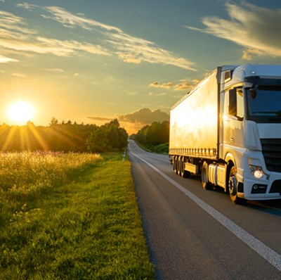 Front side view of a truck driving through a road going through grass fields with a back drop of trees a low sun and sky with white and dark blue clouds Front side view of a truck driving through a road going through grass fields with a back drop of trees a low sun and sky with white and dark blue clouds
