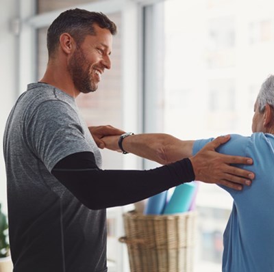 A smiling man holding the hand of a senior mans outstretched arm with one hand and the other hand on the senior mans shoulder A smiling man holding the hand of a senior mans outstretched arm with one hand and the other hand on the senior mans shoulder