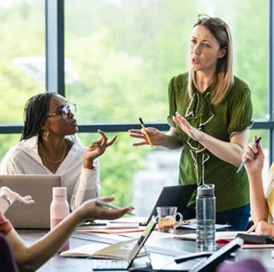 Women around a table talking with laptops, note books and bottles of water, one standing, a view of blurred greenery through floor to ceiling windows Women around a table talking with laptops, note books and bottles of water, one standing, a view of blurred greenery through floor to ceiling windows