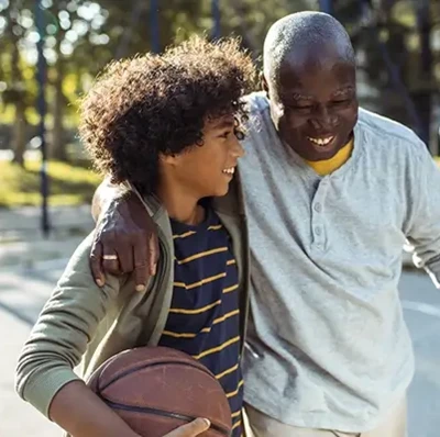 Older man smiling in a basket ball court with his arms around the shoulders of a young boy who is smiling holding a basket ball and looking at the man