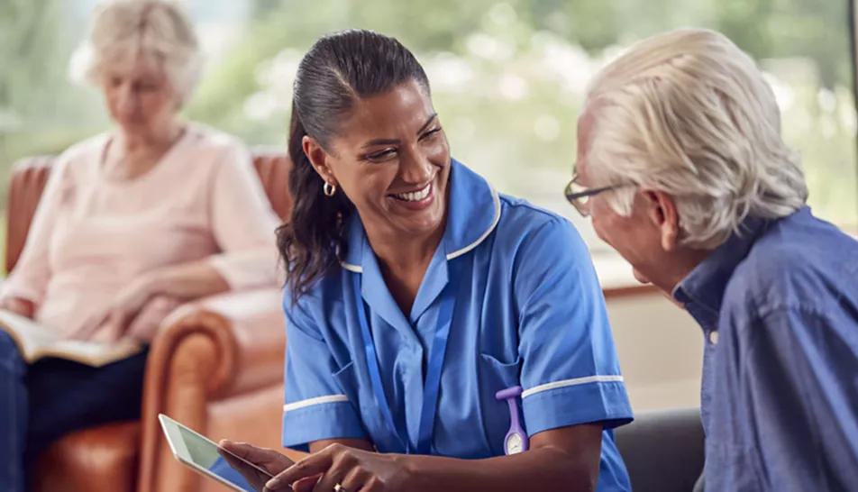 Carer smiling at elderly patient with a lady sitting in the background reading a book