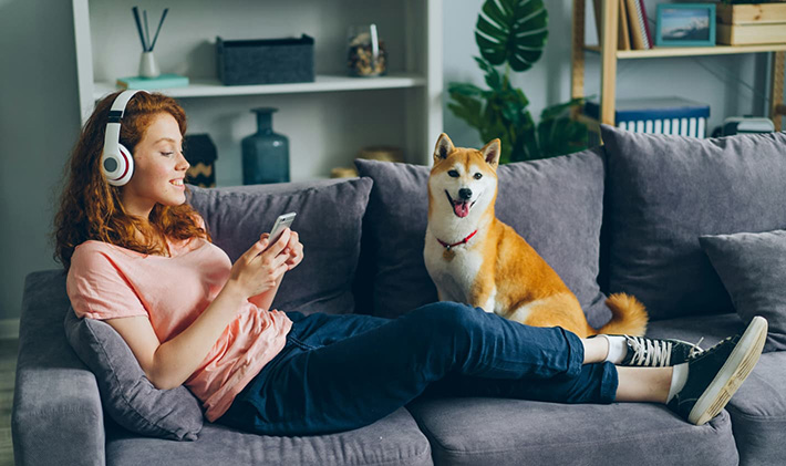Women Wearing Headphones Looking At Her Mobile With Feet Up On A Sofa with a Shiba Inu Dog Beside Her