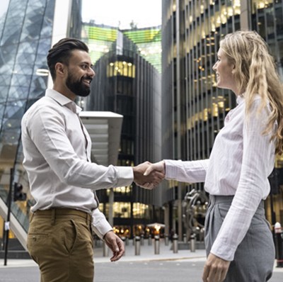 Man and a women in smart collared shirts and trousers shaking hands out side buildings in the financial district of London