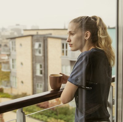 Women on a fourth floor balcony with a mug in hand looking out over modern housing estate with blurred city scape