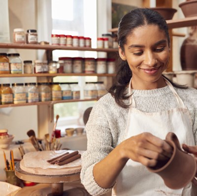 Women wearing an apron holding a mug, with a potters wheel and brushes behind her and shelves of pottery mugs, dishes, water pots and powdered glazes Women wearing an apron holding a mug, with a potters wheel and brushes behind her and shelves of pottery mugs, dishes, water pots and powdered glazes