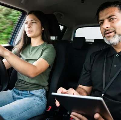 Seated view of a young adult women driving with a man next to her holding a clip board