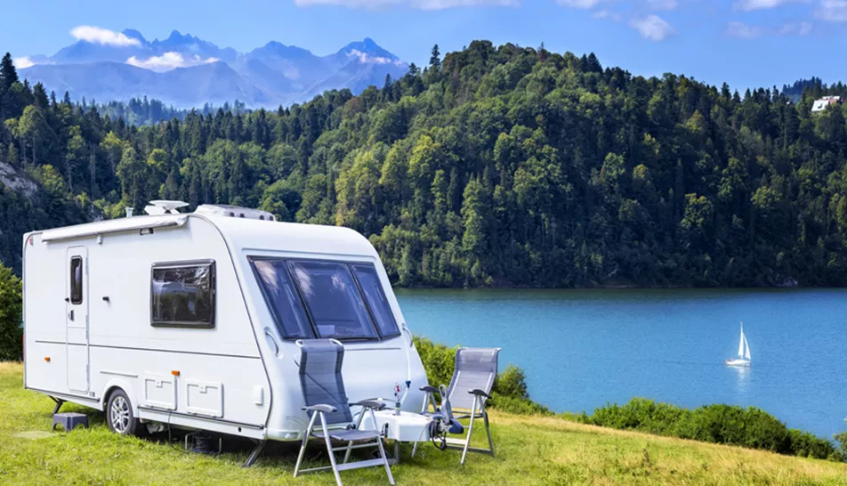 White caravan with deck chairs outside it beside a scenic lake with forest in the distance