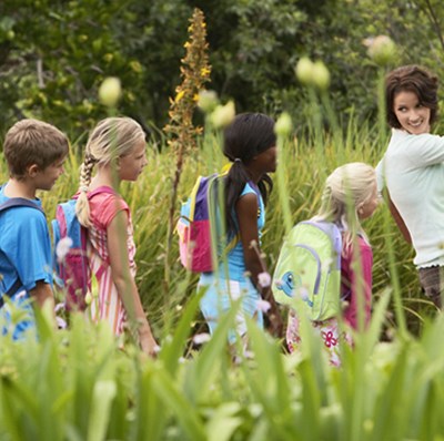 A row of children with rucksacks in a garden, looking at a woman at the front of their row, who is pointing ahead whilst looking back them and smiling
