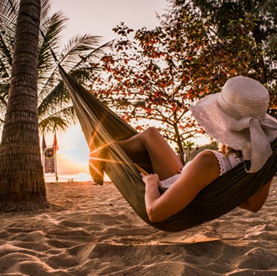 Women in a hammock on a sandy beach reading, with trees and palm trees, facing the sea on the horizon Women in a hammock on a sandy beach reading, with trees and palm trees, facing the sea on the horizon
