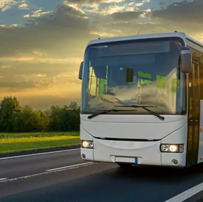 Front and side view of a coach as it drives down a road with a back drop of grass, trees, clouds and a sun low in the sky