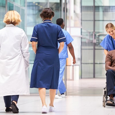 Man in a wheel chair being pushed by a wome in scrubs smiling at people in hospital uniforms in a glass lined corridor