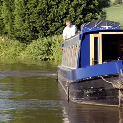 Front view of a narrow boat cruising down water with grass on the bank and a cottage in the background