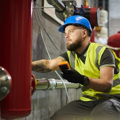 Man in a hard hat and high viz holding a small digital device, working on a large metal object in a basement setting with pipes and canisters