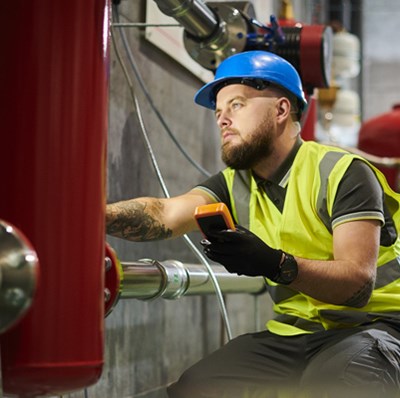 Man in a hard hat and high viz holding a small digital device, working on a large metal object in a basement setting with pipes and canisters Man in a hard hat and high viz holding a small digital device, working on a large metal object in a basement setting with pipes and canisters